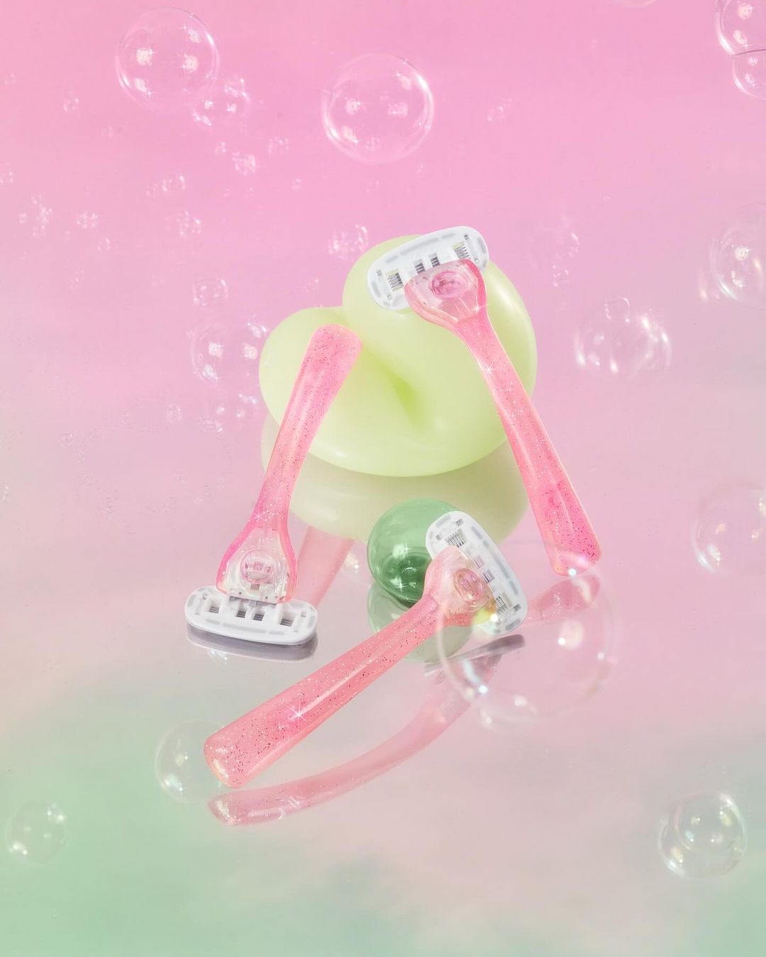 Three pink razors are arranged on a reflective surface, with a bar of soap and transparent bubble effects in a pink and green gradient.