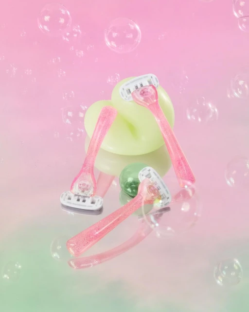 Three pink razors are arranged on a reflective surface, with a bar of soap and transparent bubble effects in a pink and green gradient.