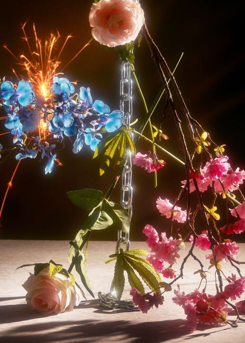 Floral still life featuring pink and blue flowers, a silver chain, green leaves, and golden sparklers against a dark background.