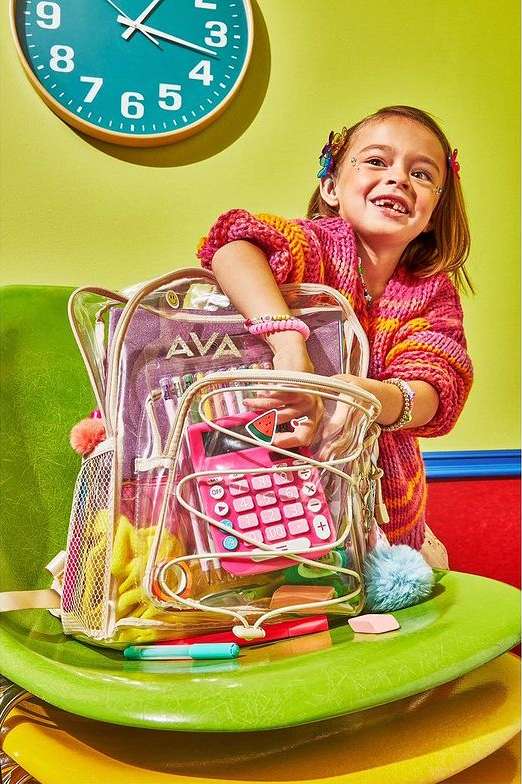 A young girl poses with a clear backpack filled with school supplies, sitting on colorful chairs against a light green wall and teal clock.