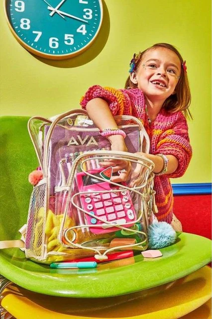 A young girl poses with a clear backpack filled with school supplies, sitting on colorful chairs against a light green wall and teal clock.