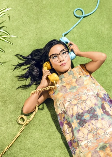 Woman with long black hair in a floral dress lying on green carpet holding retro rotary phones; one yellow, one blue.