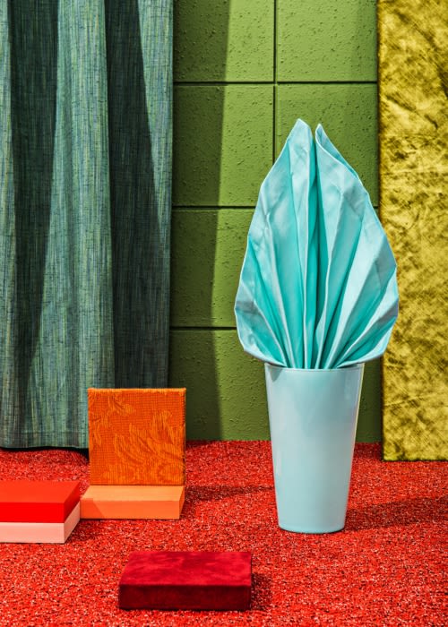Still life with blue vase holding a folded napkin, colorful cubes, and textured backgrounds in shades of green, yellow, and red.