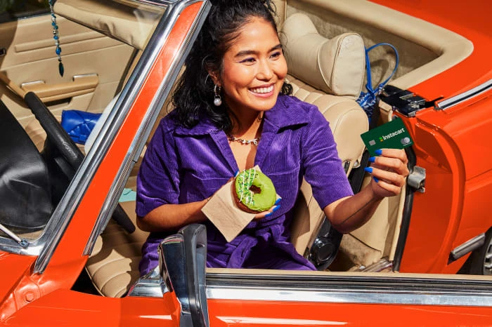 Smiling woman in a convertible car holds a green donut and an Instacart card in sunny daylight. Focus on subject and car.