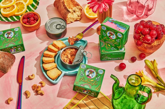 Still life of Fichusito tinned fish boxes, bread, fruit, cashews, and a glass teapot on a pink backdrop. Bright, inviting food display.