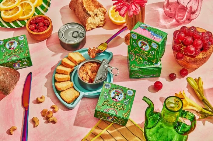 Still life of Fichusito tinned fish boxes, bread, fruit, cashews, and a glass teapot on a pink backdrop. Bright, inviting food display.