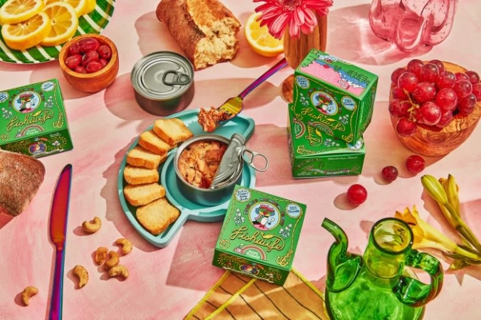 Still life of Fichusito tinned fish boxes, bread, fruit, cashews, and a glass teapot on a pink backdrop. Bright, inviting food display.