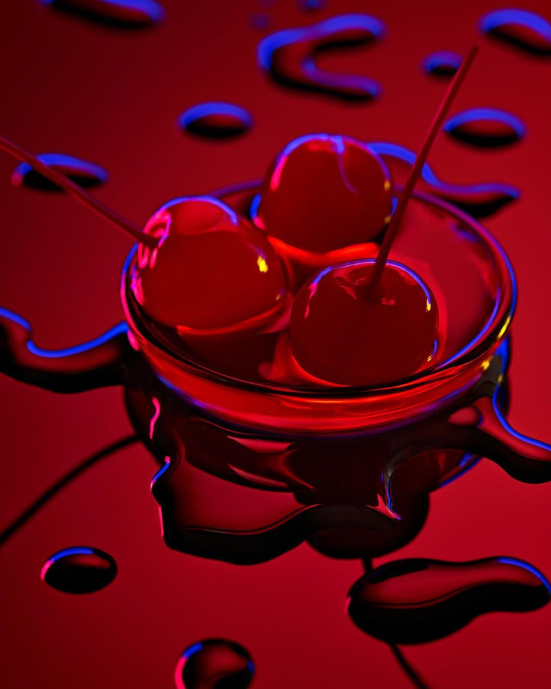 Close-up studio shot of three cherries with stems in a clear glass bowl on a glossy red surface with spilled liquid.