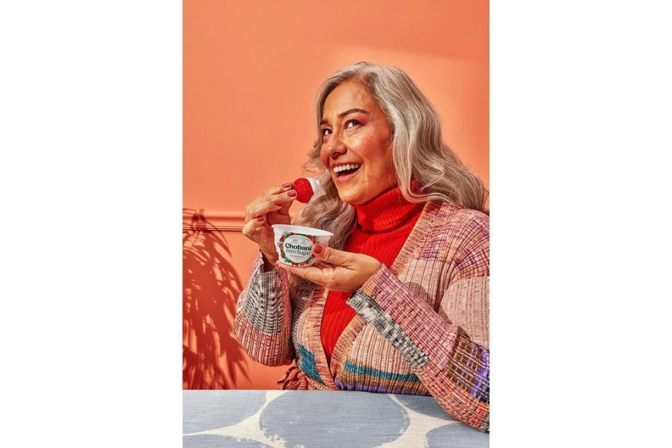 Woman in casual sweater eats strawberry with Chobani Zero Sugar yogurt against orange background in a brightly lit indoor setting.