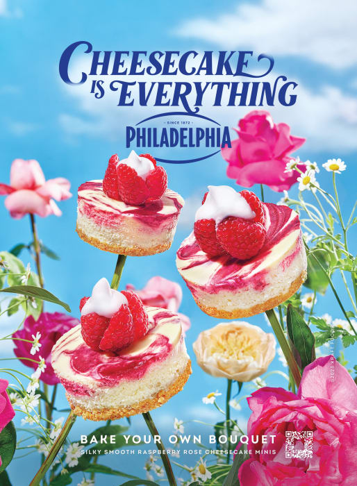Three mini cheesecakes topped with raspberries and cream float among pink and white flowers against a bright blue sky background.