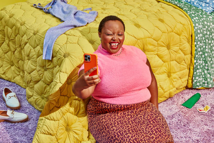 Woman in pink sweater and leopard print skirt sits on rug taking cell phone selfie with yellow quilted bed and clothing in the background.