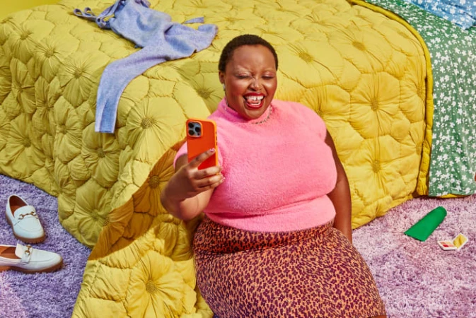 Woman in pink sweater and leopard print skirt sits on rug taking cell phone selfie with yellow quilted bed and clothing in the background.