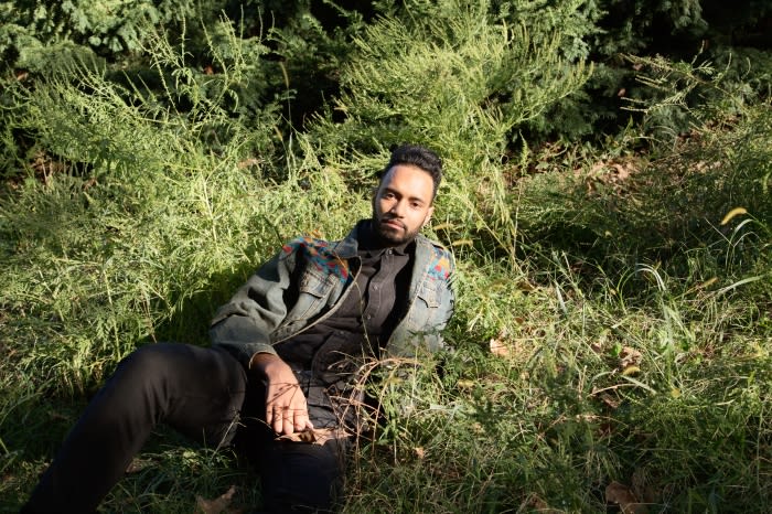 Man wearing a denim jacket seated amongst tall green foliage and grass in a natural outdoor setting