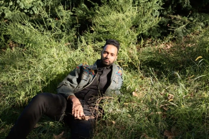 Man wearing a denim jacket seated amongst tall green foliage and grass in a natural outdoor setting