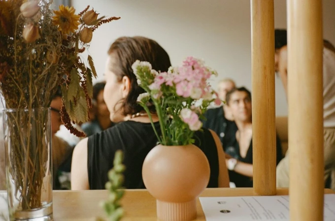 Woman in black top seated at table with pink flowers in vase and dried flowers in foreground by Naima Green