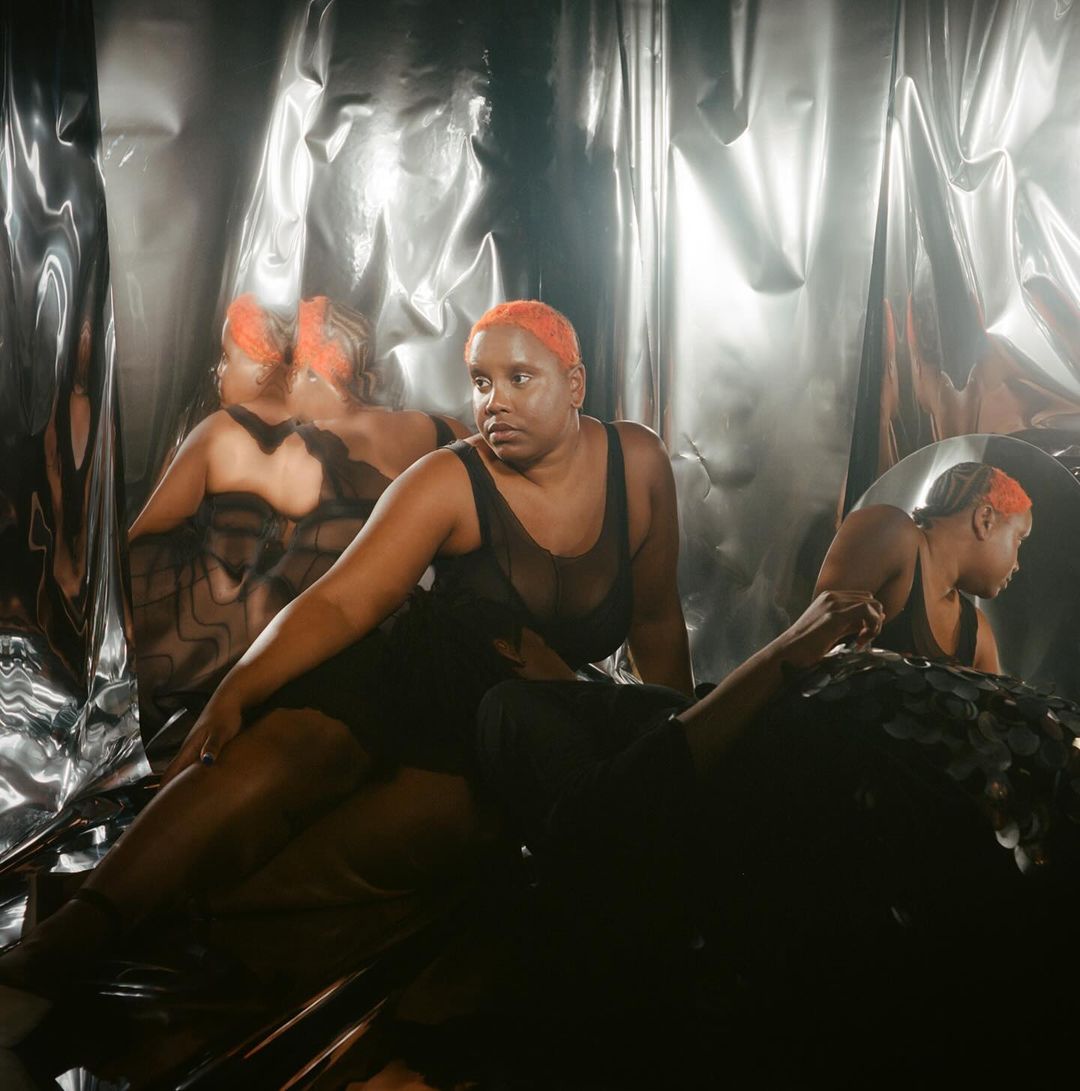 Adult woman with orange hair poses in a low-cut dress against a silver backdrop with reflections, creating a mysterious, artistic visual.