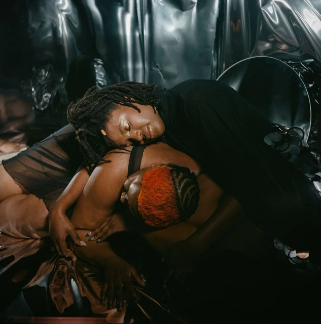 Two Black plus-size women pose intimately against a textured silver backdrop, one with short red hair, the other with dreadlocks.