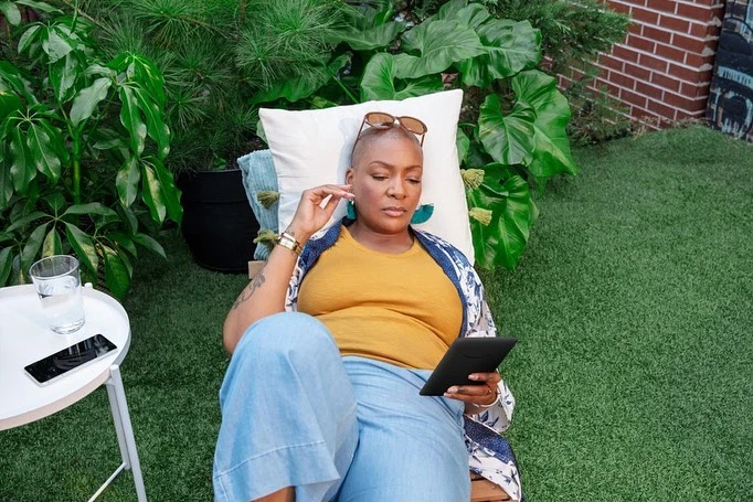 A woman with a shaved head relaxes outdoors on a pillow while looking at an electronic tablet, surrounded by green plants.
