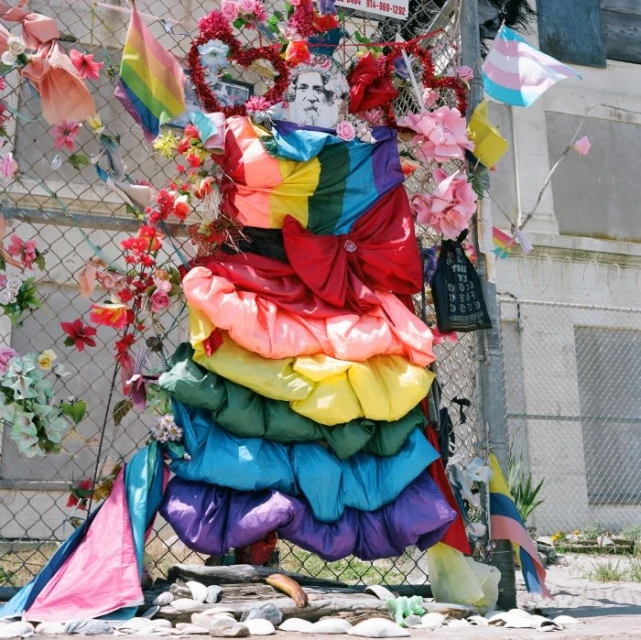 Colorful rainbow dress-like object displayed on a metal fence alongside pride flags and flowers, creating a vibrant memorial or tribute.