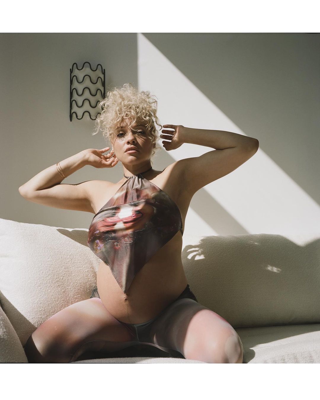 Portrait of a pregnant adult female with curly hair, wearing a printed halter top, posing indoors on a light-colored sofa in natural light.