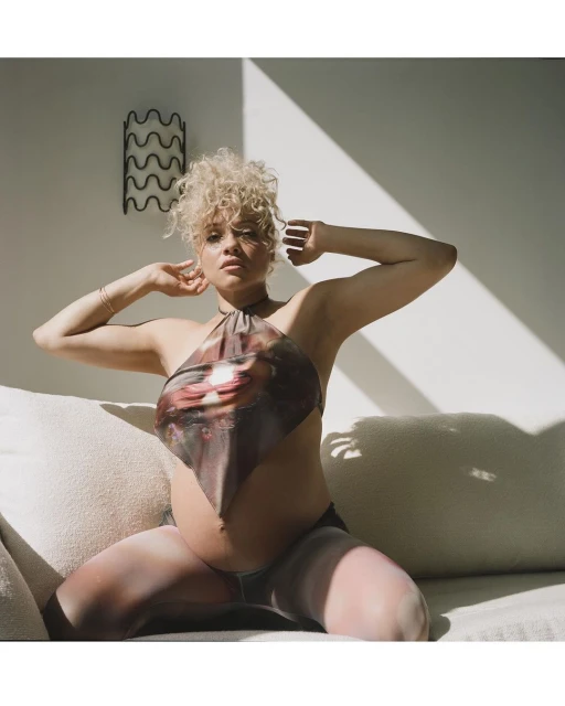 Portrait of a pregnant adult female with curly hair, wearing a printed halter top, posing indoors on a light-colored sofa in natural light.