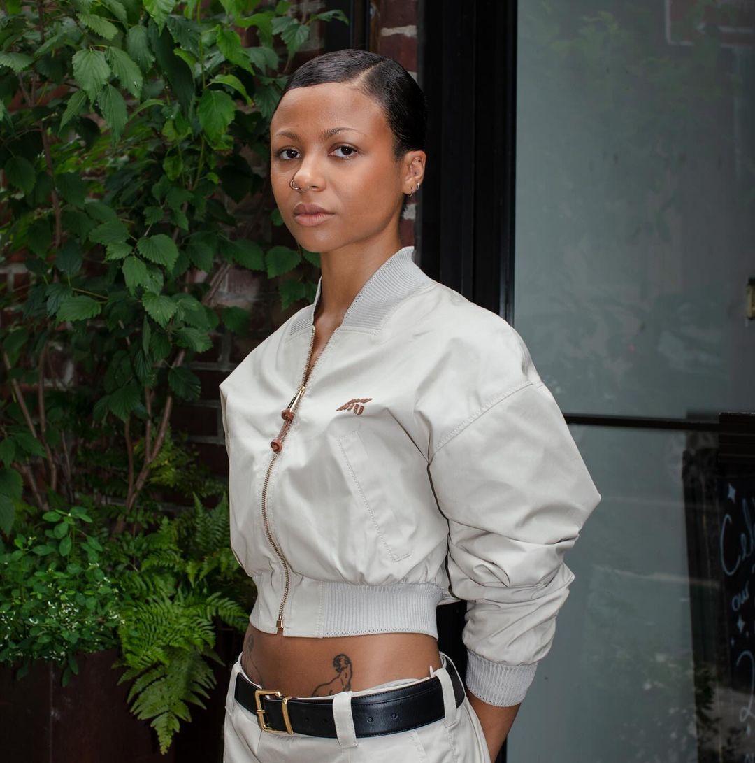 Woman in tan bomber jacket and pants poses outdoors against a brick wall with green foliage. She has a serious facial expression.