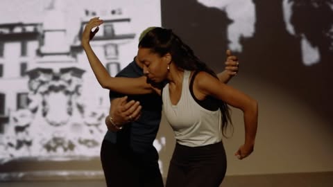 Two dancers perform in a studio with a black-and-white building projection, one in dark and the other in light workout clothes.