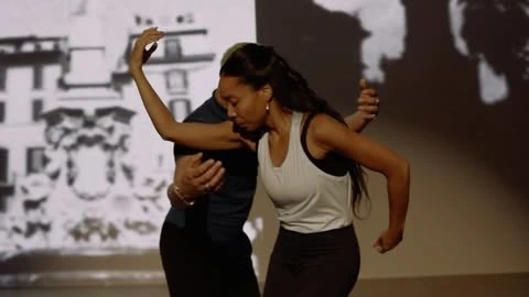 Two dancers perform in a studio with a black-and-white building projection, one in dark and the other in light workout clothes.