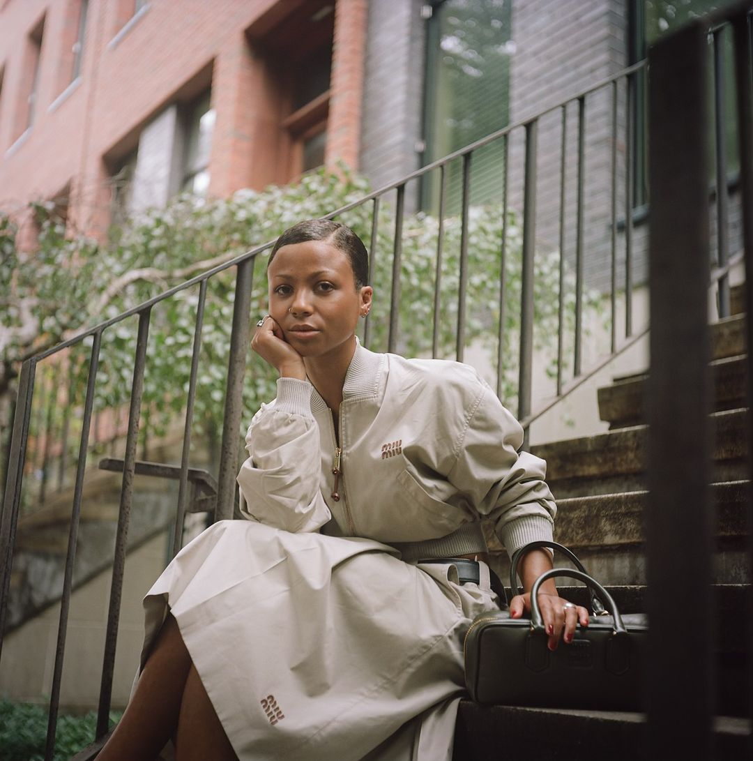 Woman wearing cream bomber jacket and skirt seated on outdoor stairs by Naima Green