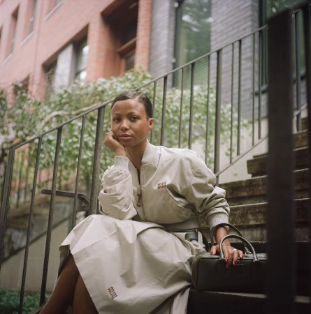 Woman wearing cream bomber jacket and skirt seated on outdoor stairs by Naima Green