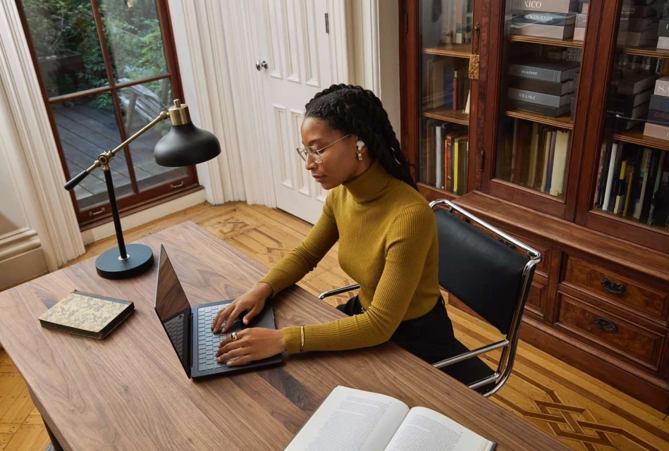 Adult female works on a laptop computer at a wooden desk in a home office with a bookcase and natural light from a window.