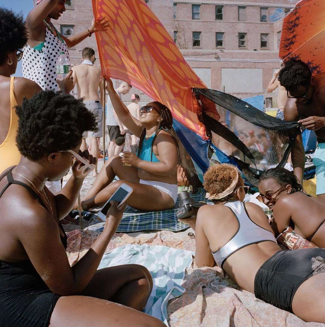 A group of young adult African American women are relaxing on a sandy beach, dressed in swimwear, with a brick building in the background.