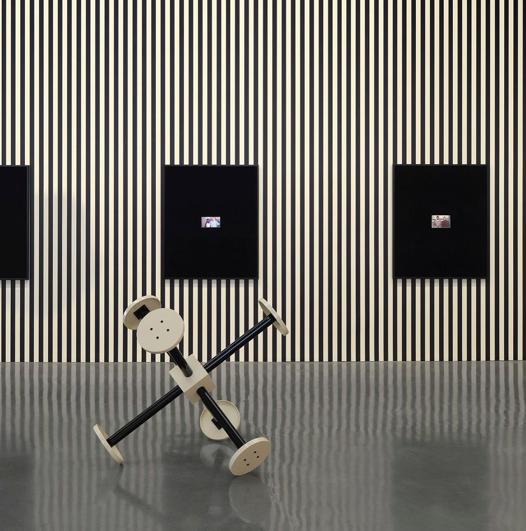 Modern art installation: abstract geometric sculpture in front of black and white striped wall with small framed artworks on display.