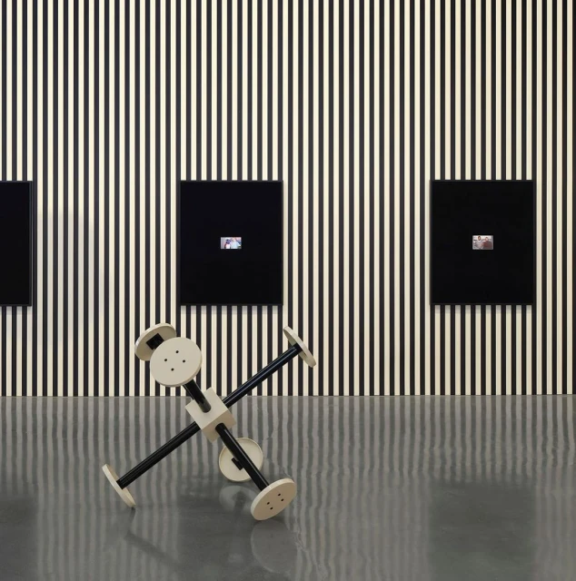 Modern art installation: abstract geometric sculpture in front of black and white striped wall with small framed artworks on display.