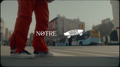 Person in orange pants and gray sneakers standing on a crosswalk with buildings in the background on an overcast day in an urban area.