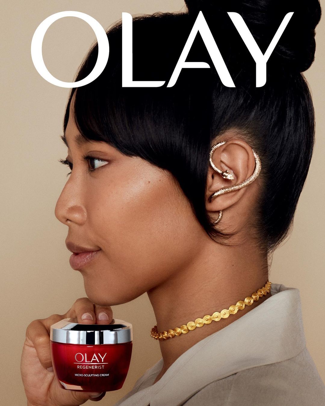 A woman poses in a close-up beauty shot with a red jar of Olay Regenerist cream, accessorized with gold jewelry against a beige background.