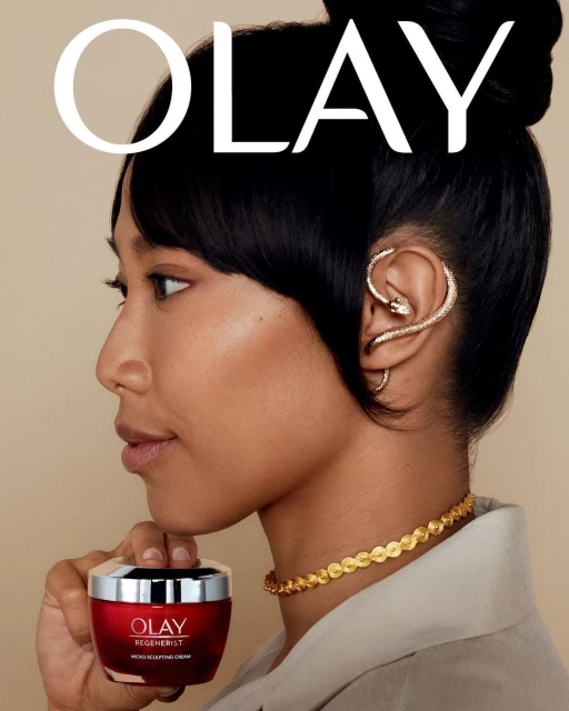 A woman poses in a close-up beauty shot with a red jar of Olay Regenerist cream, accessorized with gold jewelry against a beige background.