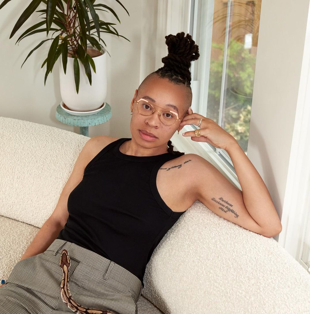 Indoor portrait of an adult woman with dark hair in a high bun, wearing glasses, a tank top, and patterned pants, on a cream couch.
