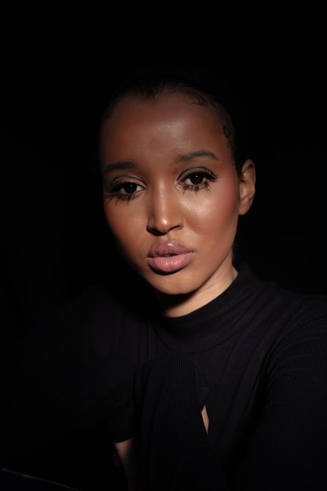 Close-up portrait of a young woman with makeup, wearing a black turtleneck sweater, against a dark backdrop with studio lighting.