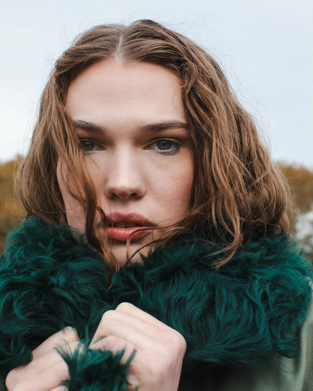 Close-up portrait of an adult woman with medium-length brown hair and striking blue eyes, wearing a faux fur collar, outdoors on a cloudy day.