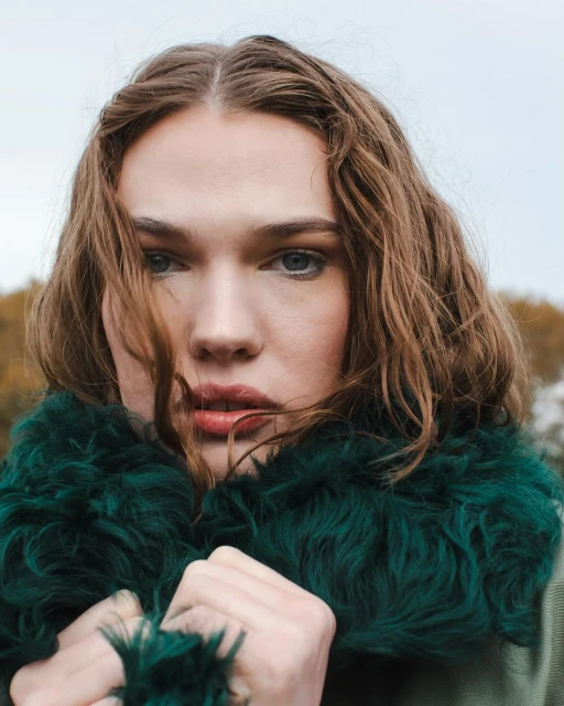 Close-up portrait of an adult woman with medium-length brown hair and striking blue eyes, wearing a faux fur collar, outdoors on a cloudy day.
