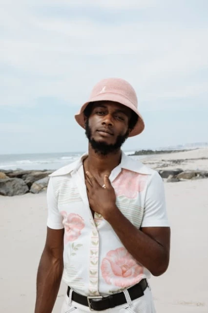 Portrait of a young Black man in a pink bucket hat and floral shirt on a beach, posing casually with the ocean and sky in the background.