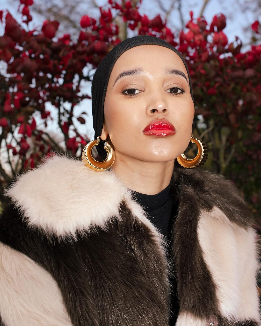 Portrait of a young adult female with fair skin, wearing a black headscarf, large gold earrings, and a faux fur coat, posing outdoors.