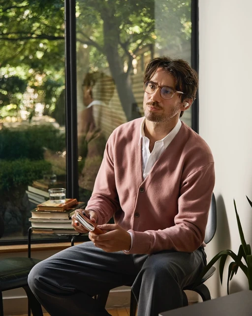 Adult man wearing glasses and a pink cardigan sitting indoors near a window with trees in the background, holding a small object.