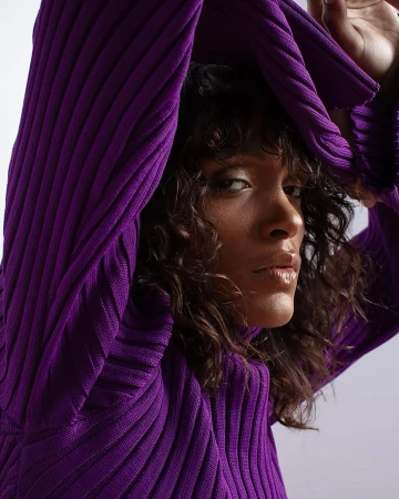 Portrait of a woman with dark curly hair wearing a ribbed purple turtleneck sweater against a white background.