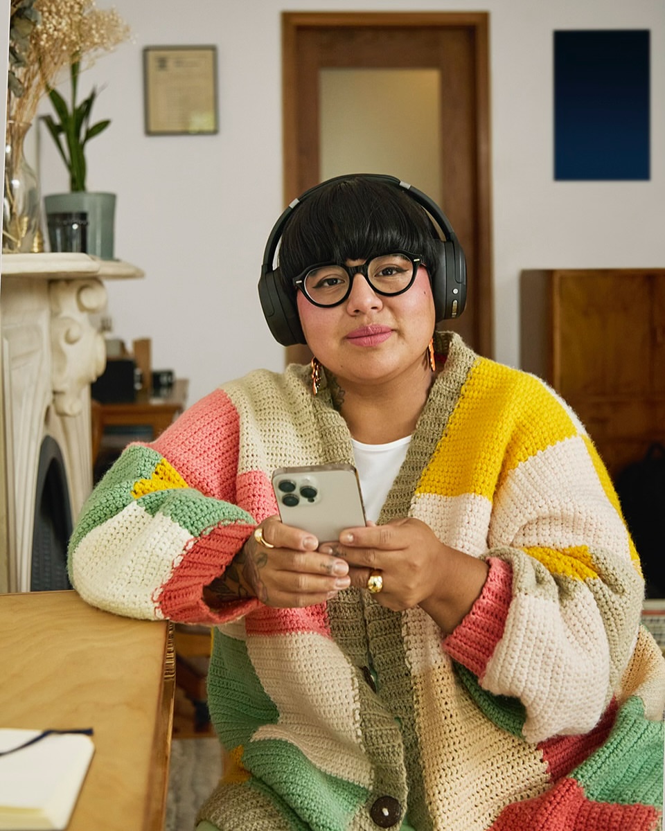 Woman wearing headphones and a colourful crochet cardigan holds a smartphone indoors with a potted plant and wall art in the background.