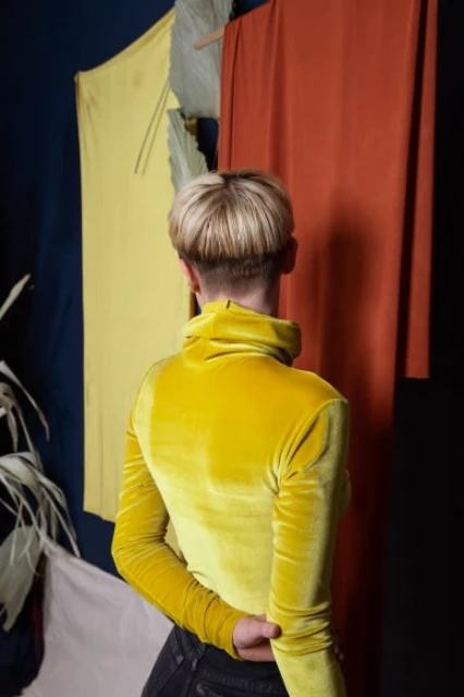 Back view of person with short blonde hair wearing a yellow turtleneck top in front of yellow and orange fabric panels.