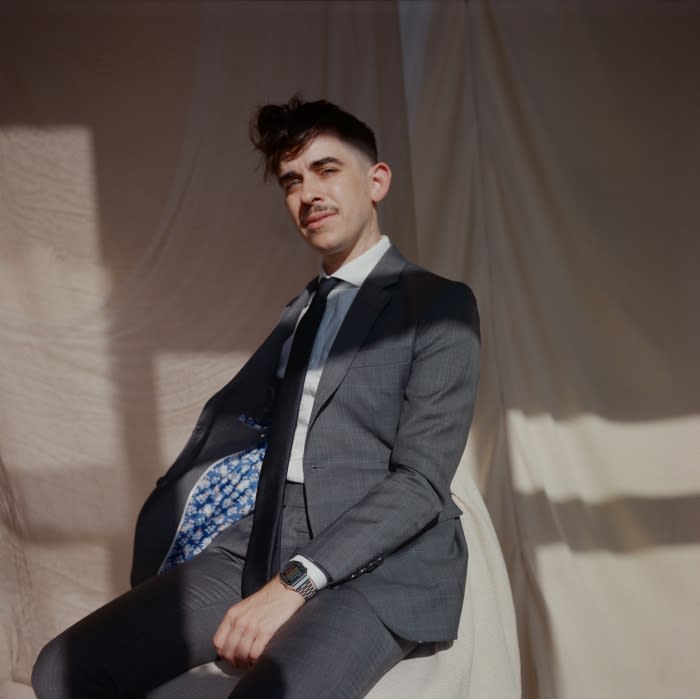 Portrait of a young adult man with a mustache wearing a gray suit, tie, and watch, sitting in front of a draped off-white backdrop.
