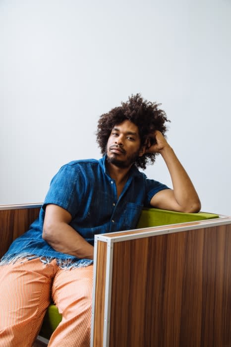 Portrait of a Black man with curly hair wearing a blue denim shirt and orange patterned trousers seated in a retro armchair.