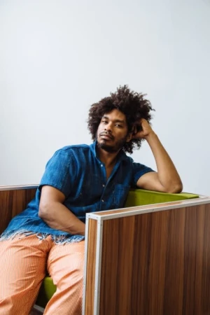 Portrait of a Black man with curly hair wearing a blue denim shirt and orange patterned trousers seated in a retro armchair.
